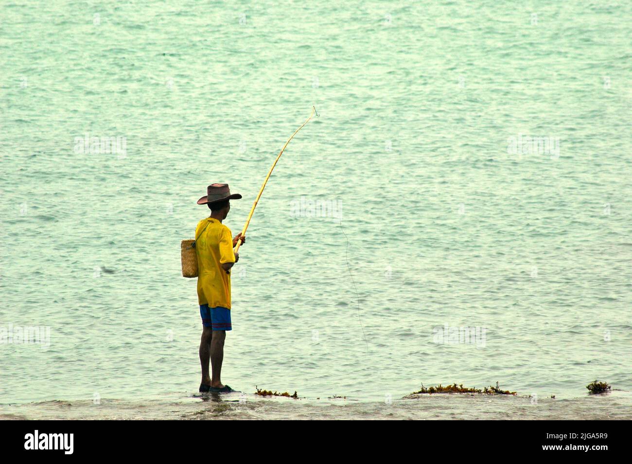 Un homme portant un chapeau de cow-boy pêche alors qu'il est debout sur des eaux peu profondes sur une plage plate et rocheuse face au détroit de Sunda à Cikoneng, Anyer, Serang, Banten, Indonésie. Banque D'Images