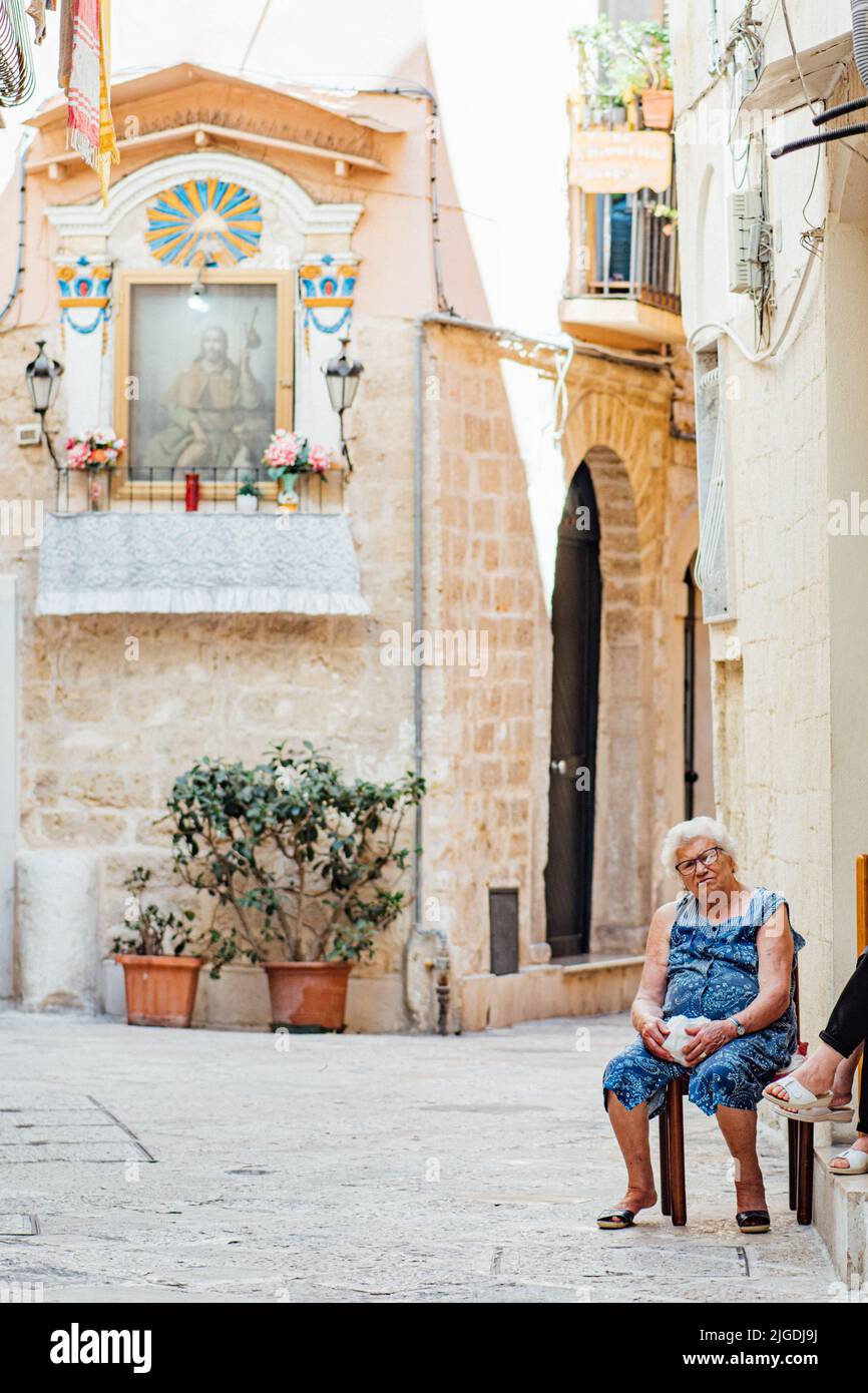 Femme âgée de la région assise sur une chaise à la porte dans une petite rue de la vieille ville de Bari, Puglia, Italie, verticale Banque D'Images