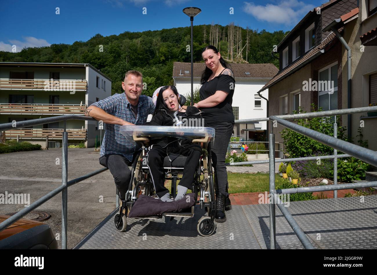 PRODUCTION - 08 juillet 2022, Rhénanie-du-Nord-Westphalie, Hagen: Dustin Raatz s'agenouille à côté du fauteuil roulant de Chantal et de sa mère Bianca Caspari devant leur maison. Raatz avait secouru la jeune fille gravement malade lors des graves inondations de l'année dernière avec un véhicule de l'armée allemande pesant plusieurs tonnes du musée et l'avait mise en sécurité après que la dernière batterie de son respirateur ait été presque vide. Ce mardi, il recevra la médaille de sauvetage de l'État du NRW par le ministre-président Hendrik Wüst (CDU) pour ses efforts courageux et altruistes. (À dpa : un conducteur de grue sauve une fille de l'inondation - avec vehi Banque D'Images
