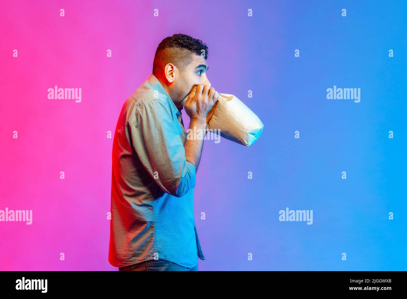 Vue latérale de l'homme nerveux dans le maillot se sentant malade, respirant dans le sac en papier pour améliorer le bien-être, surmontant le stress au travail. Studio d'intérieur isolé sur fond de néon coloré. Banque D'Images