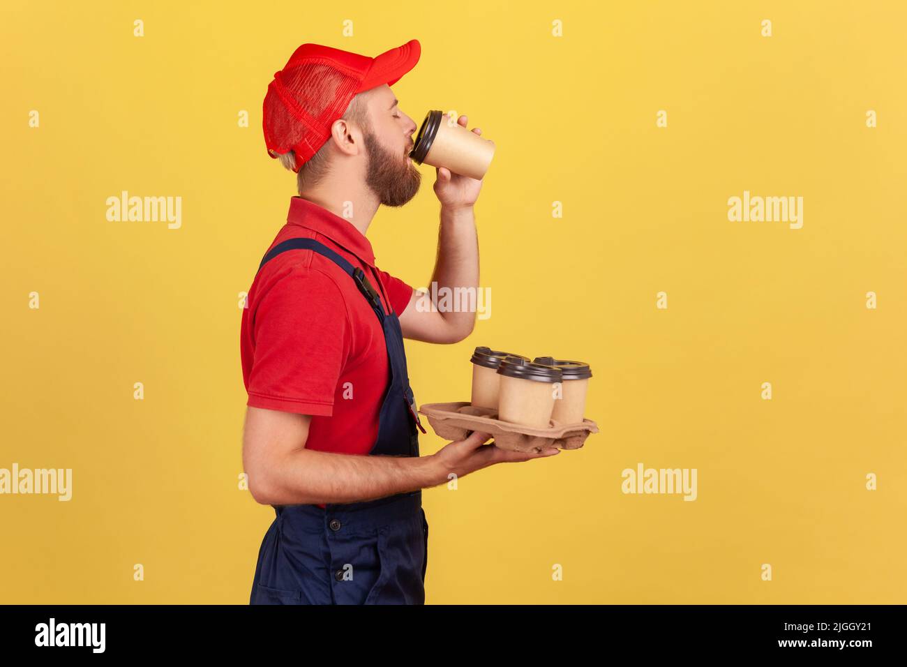 Portrait de côté de l'homme barbu maniable debout avec des tasses jetables et boire des boissons chaudes, a besoin d'énergie pour continuer à travailler. Studio d'intérieur isolé sur fond jaune. Banque D'Images