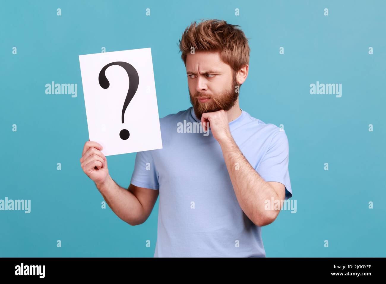 Portrait d'un homme perplexe barbu tenant un livre blanc avec un point d'interrogation, trouvant une solution intelligente, demandant des conseils, tenant le menton. Studio d'intérieur isolé sur fond bleu. Banque D'Images