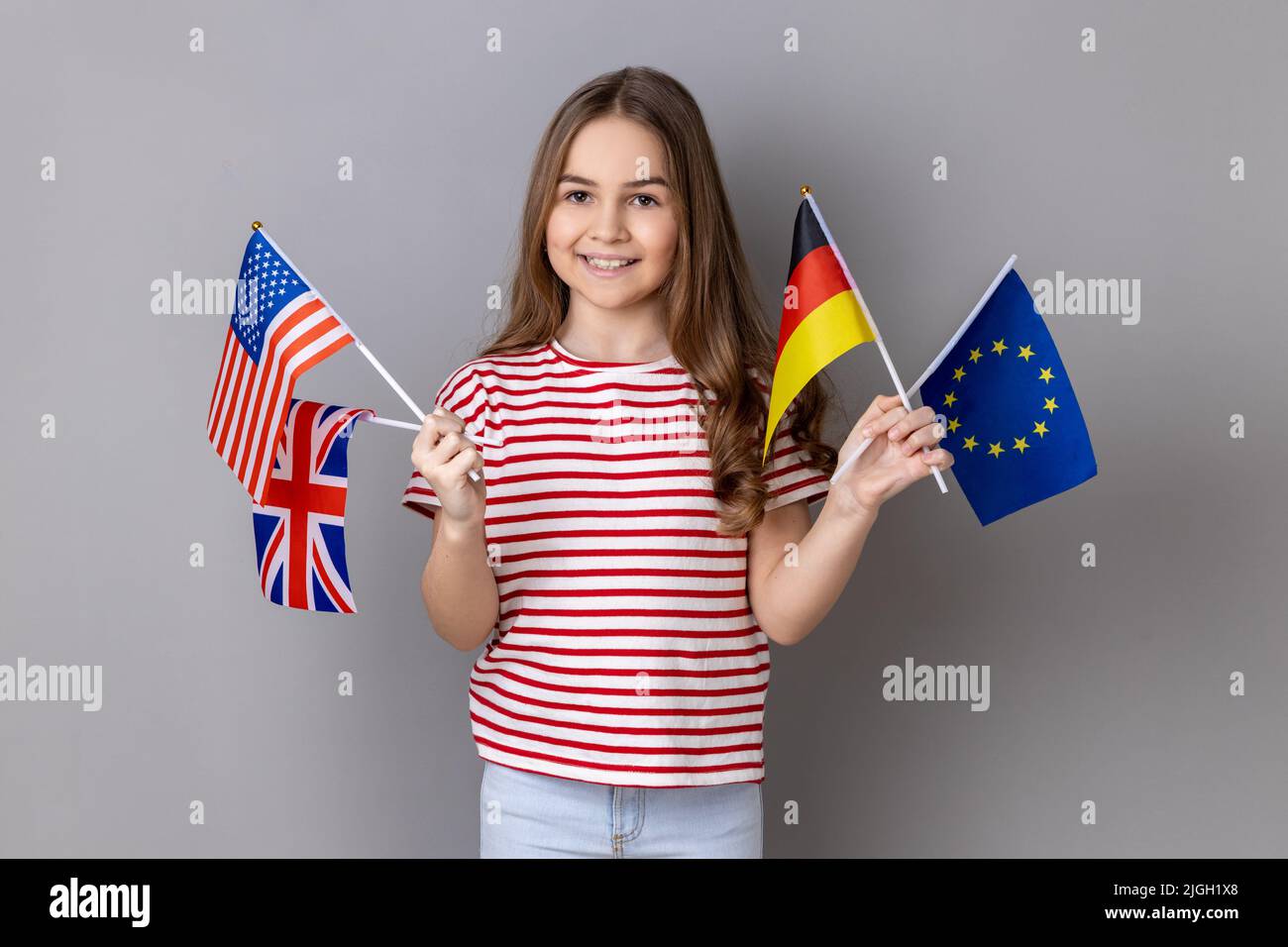 Portrait d'une petite fille adorable et optimiste portant un T-shirt rayé regardant l'appareil photo, tenant des drapeaux européens, américains, allemands et britanniques. Prise de vue en studio isolée sur fond gris. Banque D'Images