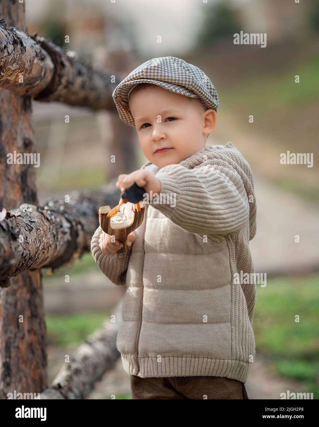 Un petit garçon intimide avec un coup de feu vise quelqu'un près d'une clôture dans le village à l'extérieur. Un enfant rustique pieds nus avec un chapeau tire une balle. Banque D'Images