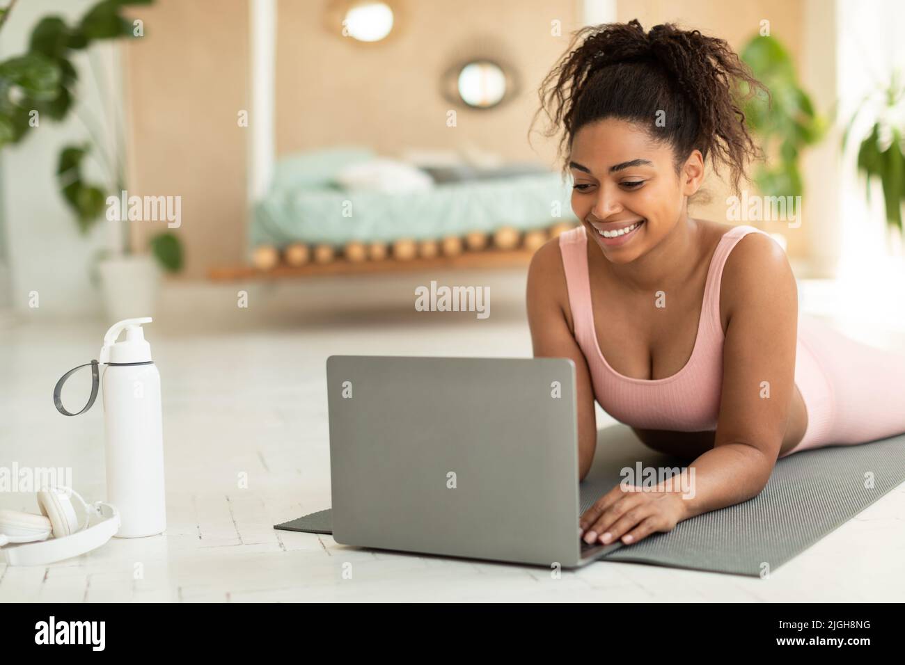 Bonne jeune femme noire couchée sur un tapis et utilisant un ordinateur portable, parlant à l'entraîneur sur le Web ou regardant des vidéos de sport Banque D'Images