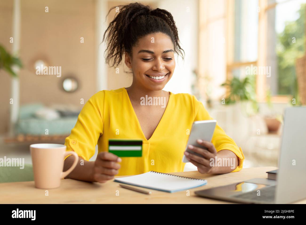 Bonne dame noire achetant des choses en ligne, en utilisant un smartphone et une carte de crédit, magasiner sur Internet tout en étant assise à la maison Banque D'Images