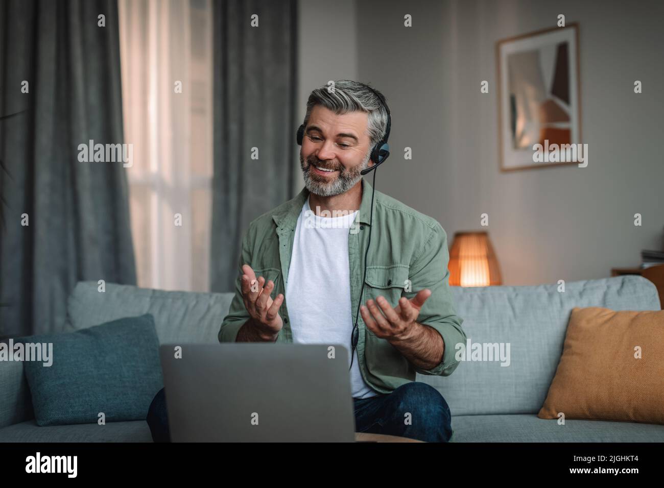 Joyeux souriant vieux caucasien avec la barbe dans le casque gestuelle, parle et regarde l'ordinateur portable Banque D'Images