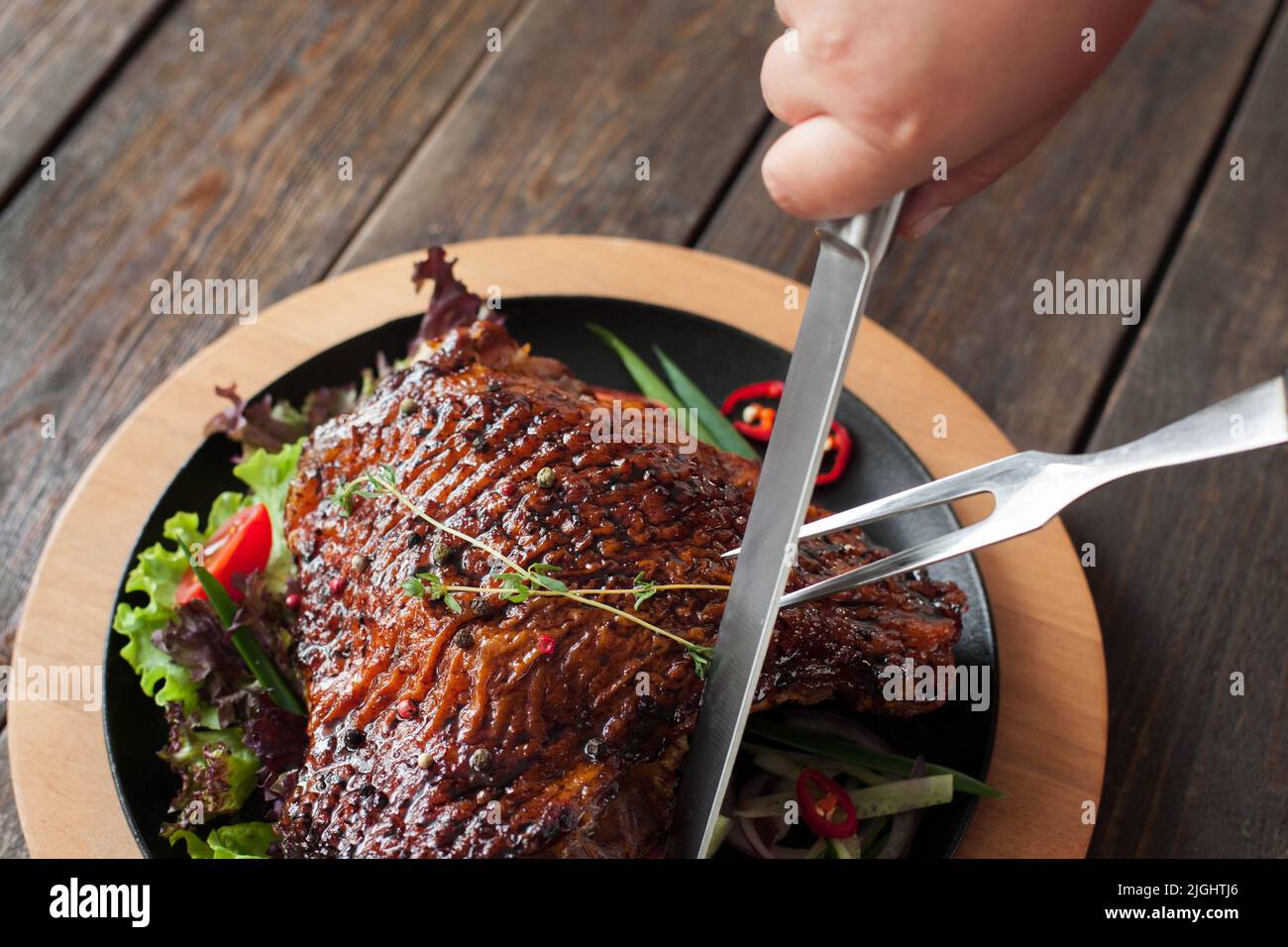 Coupe de cuisse de canard grillée vue rapprochée du dessus. Sculpter les mains de la jambe de volaille rôtie avec une fourchette à deux pattes et un couteau sur fond de bois Banque D'Images