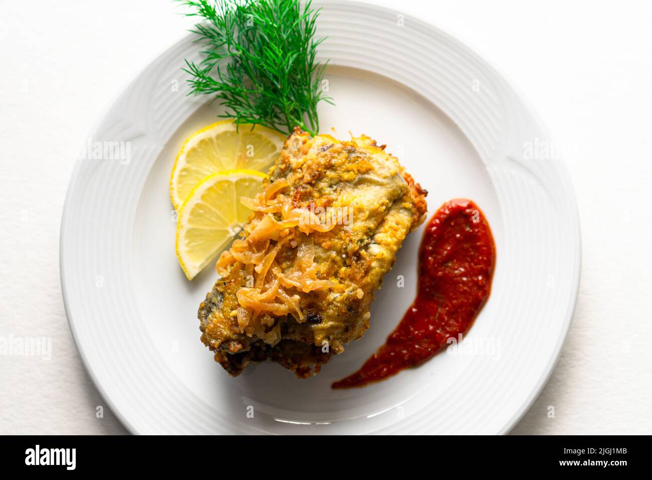 Poisson frappé sur une plaque de céramique blanche avec citron, sauce rouge et herbes sur un fond blanc isolé. Ingrédients : morue, pâte, aneth. Banque D'Images