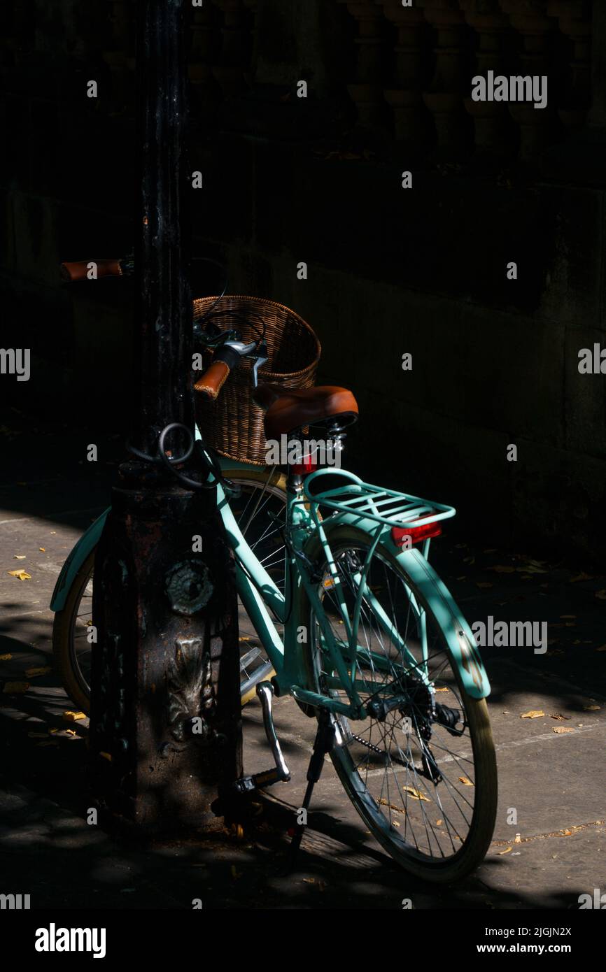 Vélo turquoise avec un panier avant enchaîné à un lampadaire sous l'ombre d'un arbre. Banque D'Images