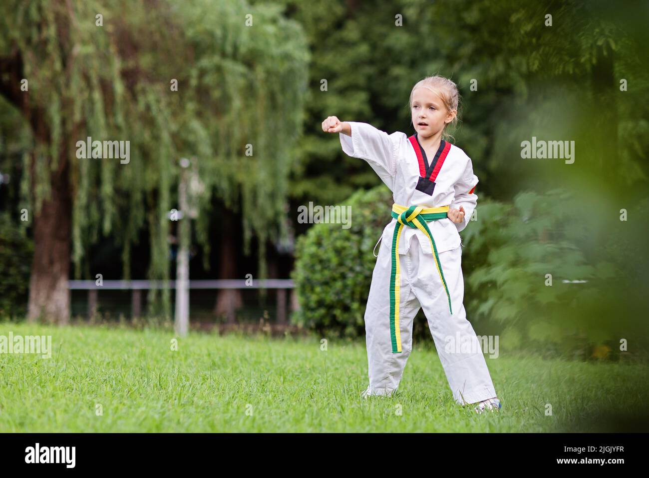 Petite fille caucasienne de sept ans dans un kimono avec ceinture verte jaune faisant de l'exercice de Taekwondo au parc d'été seul pendant le verrouillage du coronavirus covid-19 Banque D'Images