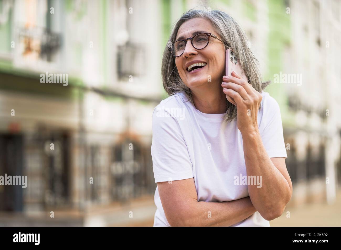 Charmante femme d'affaires mûre aux cheveux gris parlant sur le téléphone tenant un smartphone debout à l'extérieur des rues de la vieille ville urbaine portant un t-shirt blanc. Femme argentée de cheveux à l'extérieur. Banque D'Images