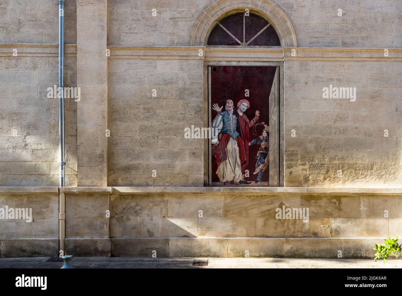 Une fenêtre peinte montre deux personnages théâtraux masqués en costume rouge et blanc-bleu avec deux enfants devant une porte rouge, sur une façade dans la vieille ville d'Avignon, en France. Depuis 1986, l'artiste Marion Pochy et l'artiste Dominique Durand ont peint environ 70 de ces fenêtres dans toute la ville avec des motifs de productions passées du Festival d'Avignon, fondé par Jean Vilar en 1947. Plusieurs de ces fenêtres ont été offertes par la société JCDecaux. Banque D'Images