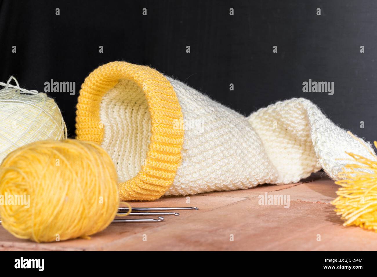 le chapeau d'elf de bébé crocheted sur des planches en bois et un fond noir, à côté de lui deux rouleaux de fil jaune et blanc, des matériaux pour commencer le crocheting. concept Banque D'Images