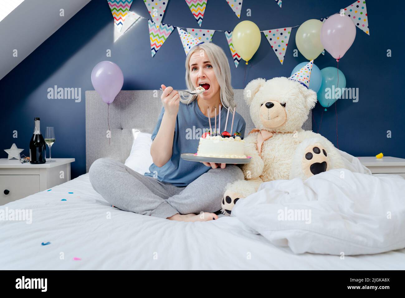 Triste, bouleversée femme en pajama et tête de fête mangeant en cuillère son gâteau d'anniversaire assis sur le lit avec un immense ours en peluche dans la chambre décorée. Fête le bi Banque D'Images