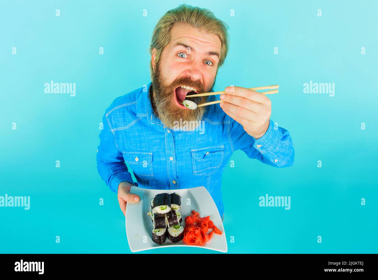 Homme barbu avec une assiette de petits pains à sushis et de gingembre mariné. Un gars mangeant un rouleau maki avec des baguettes. Banque D'Images