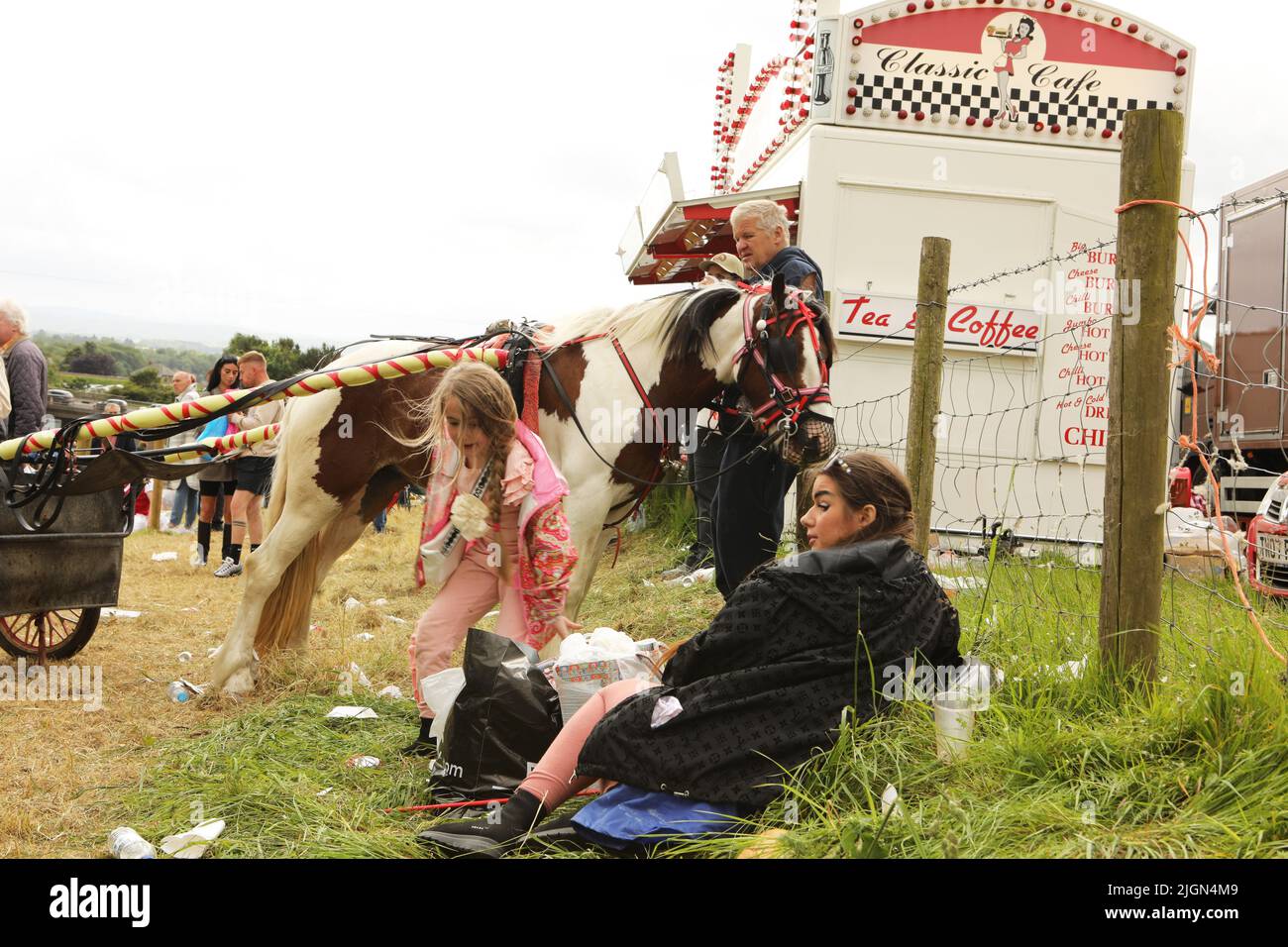 Une mère et un enfant assis sur l'herbe devant un homme debout avec un poney coloré et un piège. Appleby Horse Fair, Appleby à Westmorland, Cumbria Banque D'Images