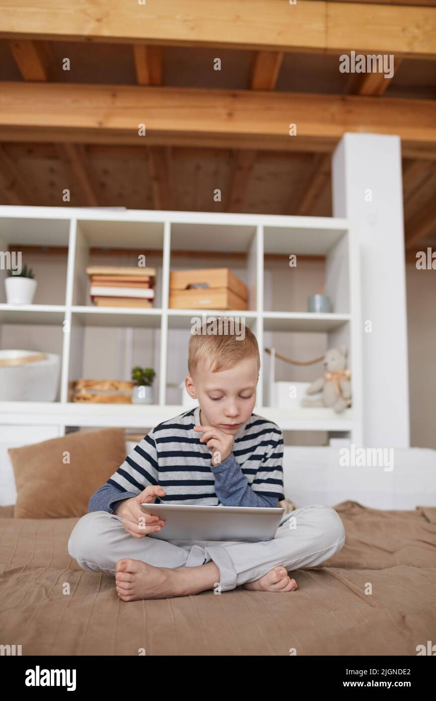 Un petit garçon sérieux et attentionné aux cheveux blonds assis avec des jambes croisées sur le lit et regardant un film éducatif sur une tablette Banque D'Images