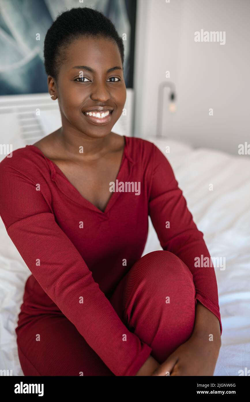 Portrait d'une jeune femme africaine souriante et heureuse dans un intérieur moderne. Fille noire assise sur le bord de son lit regardant la caméra avec le visage gai Banque D'Images