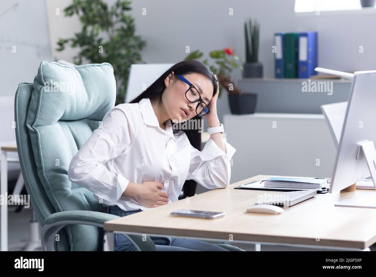 Douleur abdominale sur le lieu de travail. Une jeune belle femme asiatique en lunettes travaille dans le bureau à l'ordinateur, sent la douleur dans son estomac, tient sa main à son estomac, grimace dans la douleur. Banque D'Images