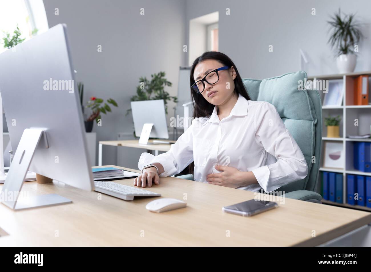 Douleur abdominale sur le lieu de travail. Une jeune belle femme asiatique en lunettes travaille dans le bureau à l'ordinateur, sent la douleur dans son estomac, tient sa main à son estomac, grimace dans la douleur. Banque D'Images