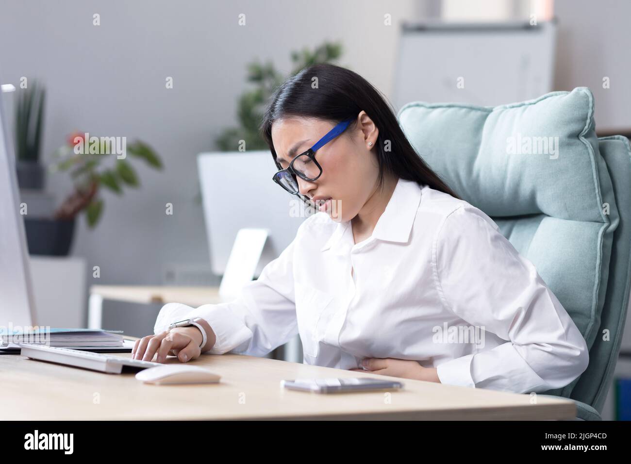 Gros plan. Douleur abdominale sur le lieu de travail.Une jeune femme asiatique en lunettes travaille dans le bureau à l'ordinateur, sent la douleur dans son estomac, tient sa main à son estomac, grimace dans la douleur. Banque D'Images