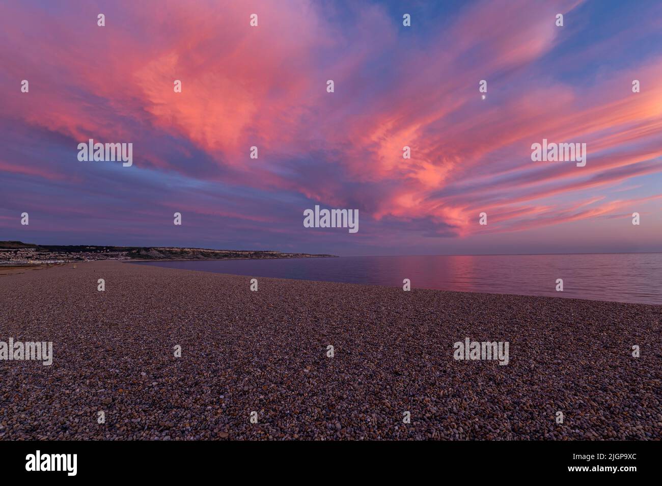 Ciel rose tandis que le soleil se couche sur la plage de Chesil avec une rive de galets et une mer calme et plate reflétant les nuages roses lumineux le long de la côte jurassique de Dorset. Banque D'Images