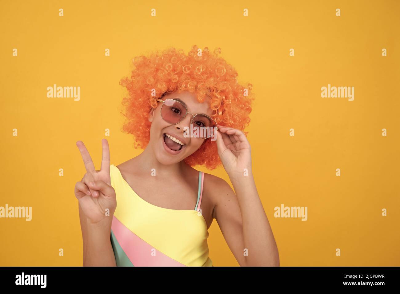 joyeux enfant dans des lunettes de soleil et un maillot de bain portant des cheveux de perruque orange montrant le geste de paix, la paix Banque D'Images