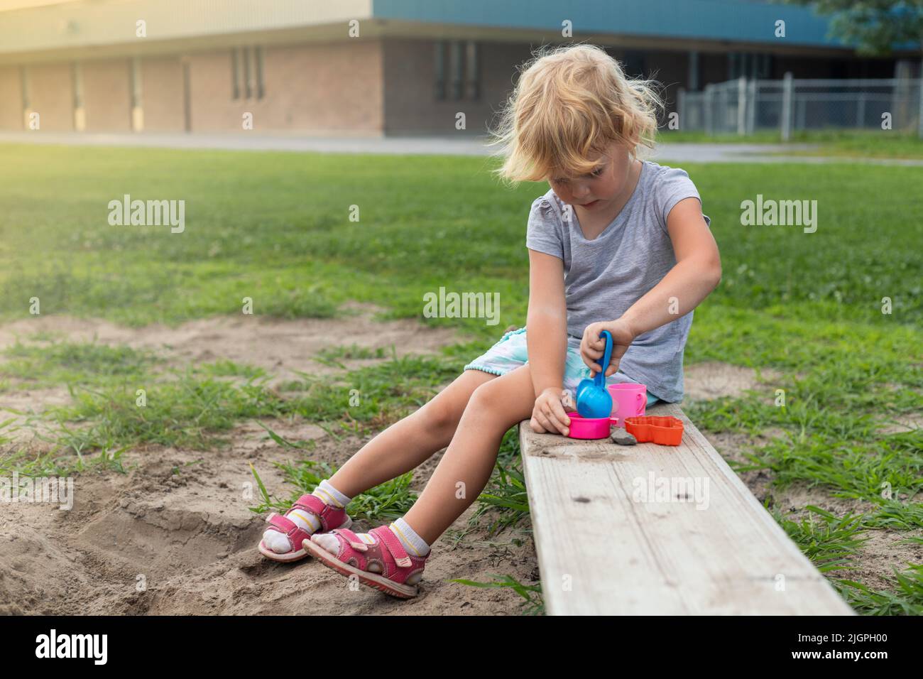 Petit enfant jouant avec du sable et des jouets à l'extérieur dans la cour d'école le jour ensoleillé. Petite fille assise sur le banc à l'aire de jeux en été Banque D'Images