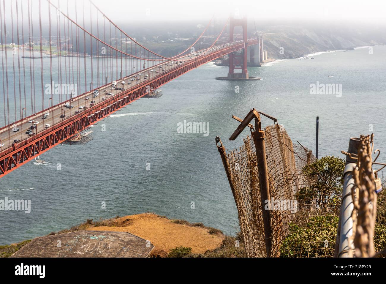 Le Golden Gate Bridge s'est tenu en hauteur, capturant une clôture en ruine avec des détails atmosphériques tandis qu'un bateau à voile passe en dessous pendant un après-midi d'été. Banque D'Images