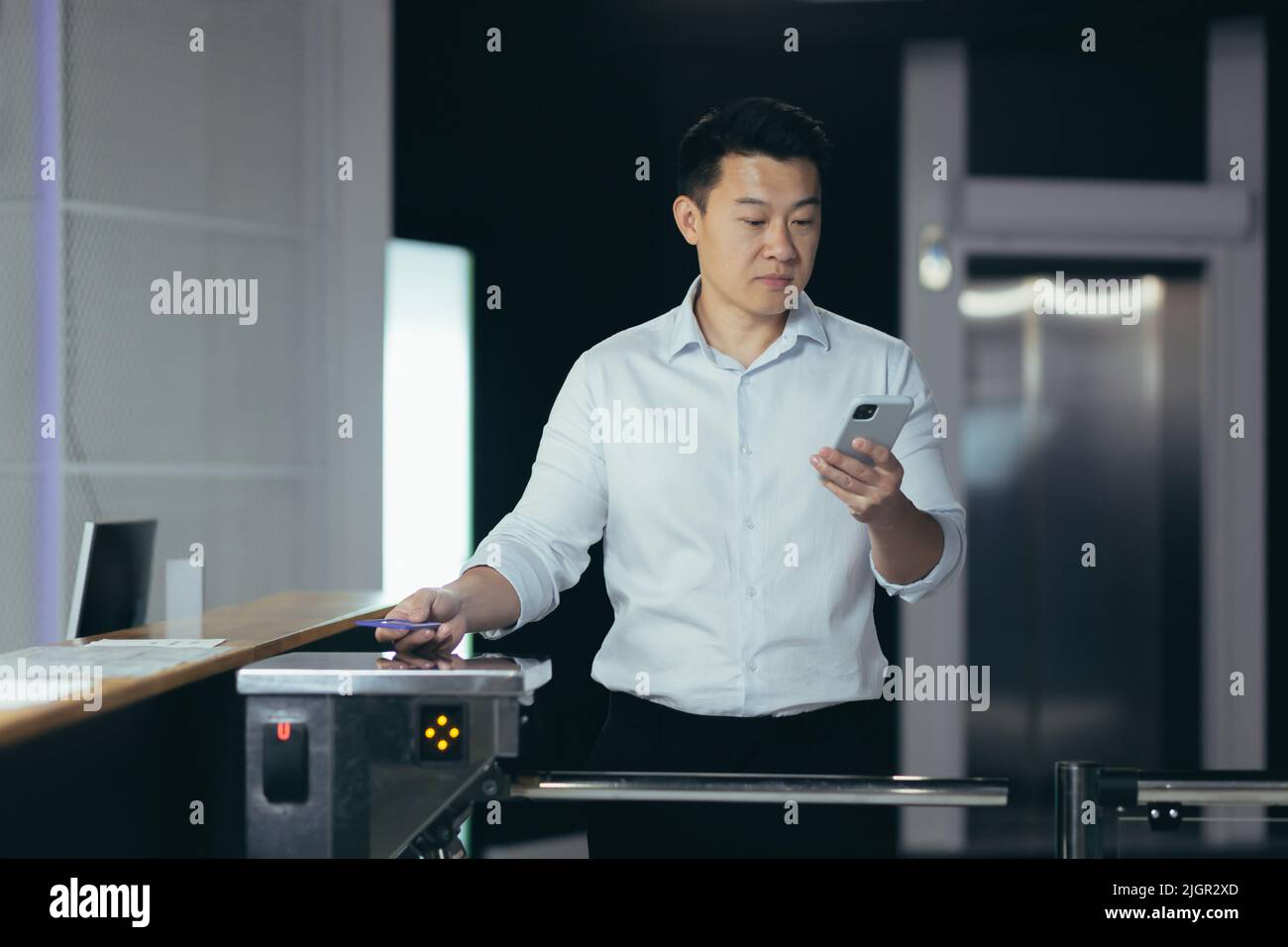Un homme d'affaires asiatique prospère entre au bureau, utilise la carte-clé pour ouvrir le tourniquet, l'homme regarde l'écran du téléphone lit les nouvelles Banque D'Images