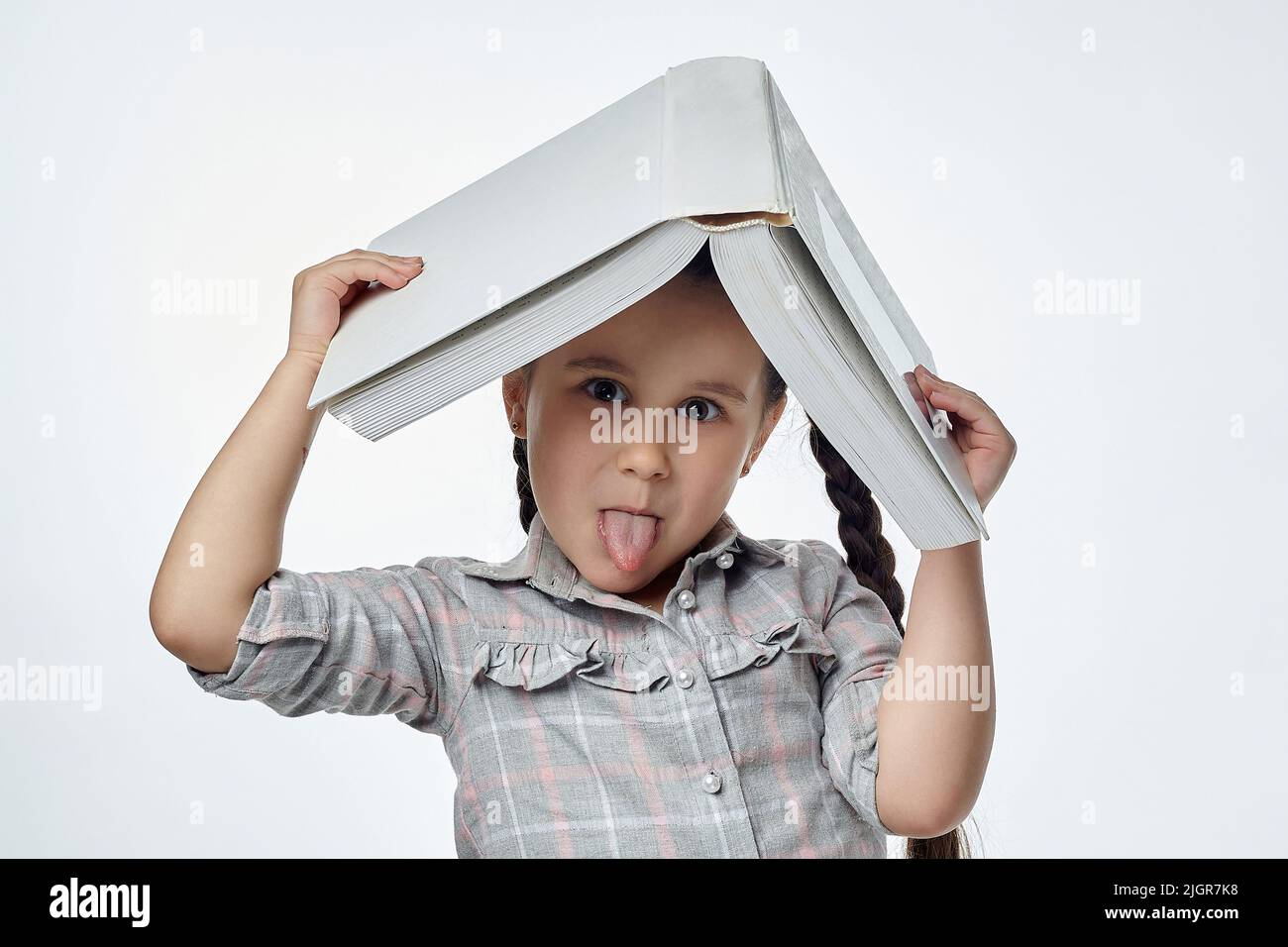 petite fille souriante avec des picots tressés tient un grand livre au-dessus de sa tête et montre sa langue. Banque D'Images