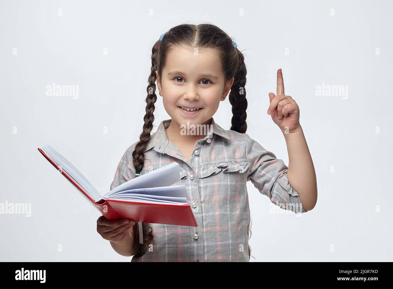 une petite fille à cheveux noirs tient un livre rouge dans ses mains et pointe vers l'espace libre avec son index. Banque D'Images