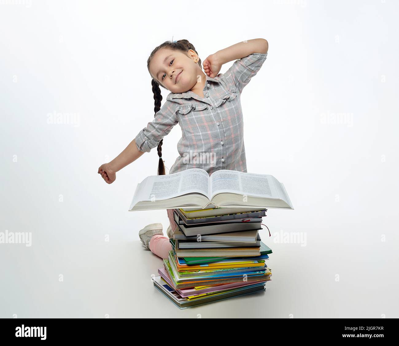 une petite fille fatiguée est assise devant une grande pile de livres et s'étire. Banque D'Images