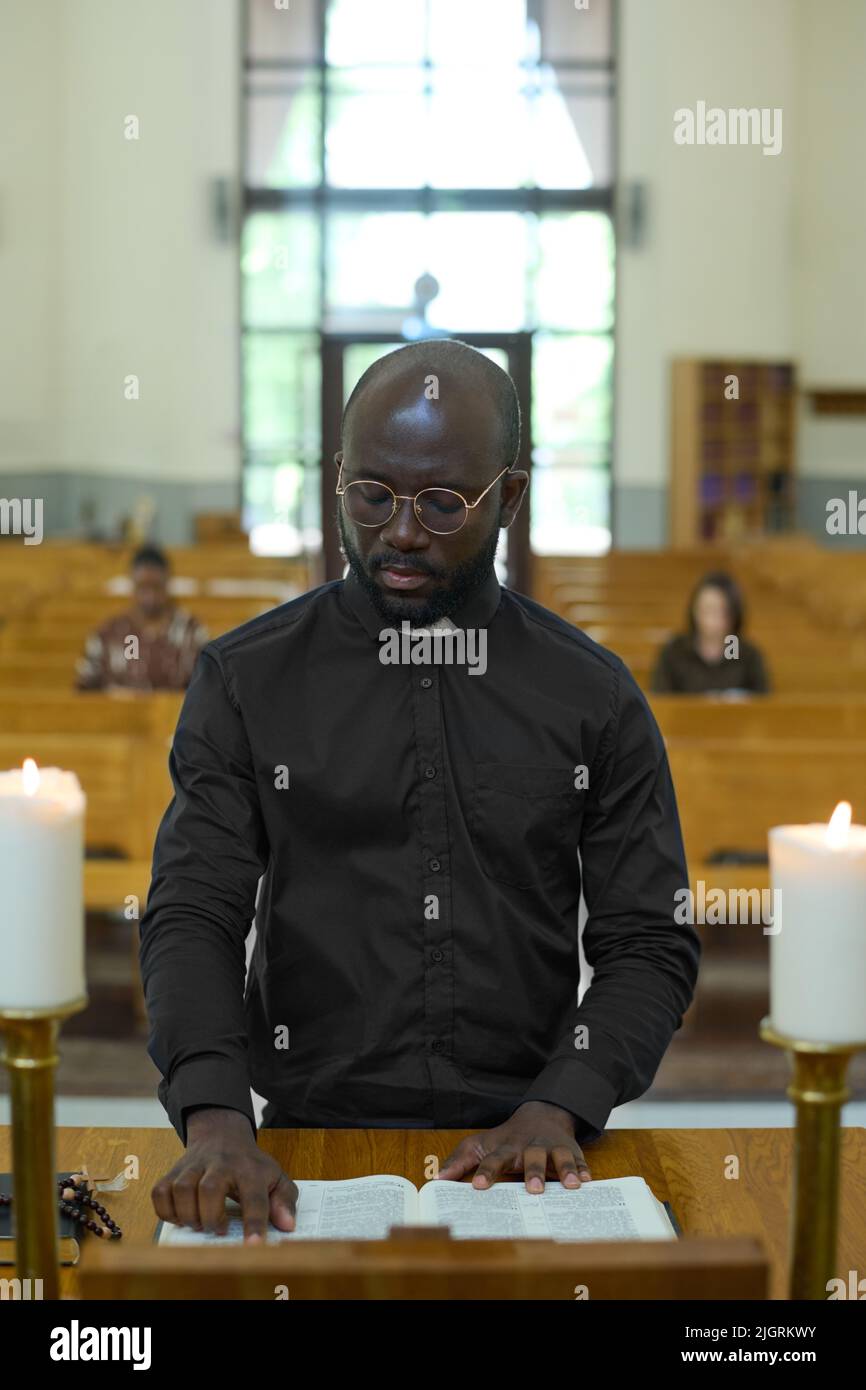 Jeune Africain américain en chemise noire avec collier clérical lisant un des quatre Evangiles ou un autre livre de la Sainte Bible pendant la liturgie Banque D'Images