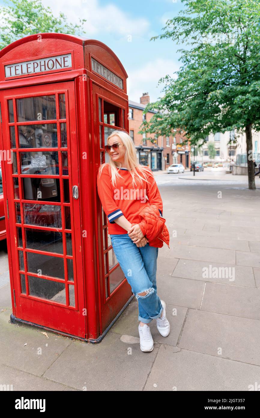 La femme heureuse portant un Jean et une chemise rouge a une date contre la boîte téléphonique rouge dans la ville anglaise Banque D'Images