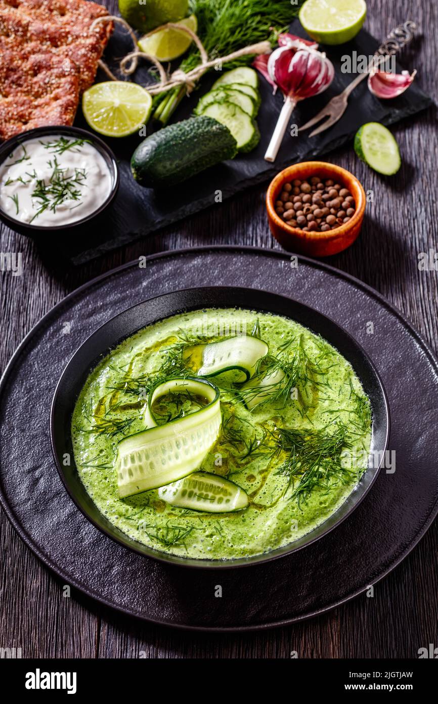 Tarator, soupe d'été au concombre froid, avec aneth, yaourt, huile d'olive dans un bol noir sur table en bois foncé avec ingrédients et pain de seigle, cuis méditerranéen Banque D'Images
