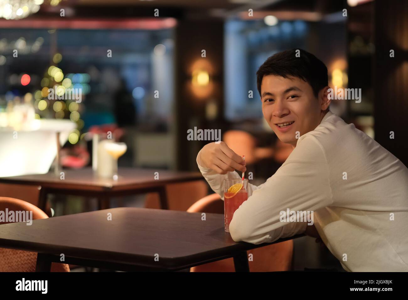 Un jeune homme asiatique souriant boit un cocktail à la table du bar, se retournant et regardant la caméra la nuit Banque D'Images