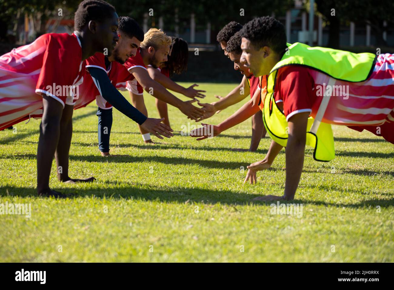 Joueurs masculins multiraciaux en uniforme rouge pratiquant cinq push-up dans une rangée sur un terrain herbeux Banque D'Images