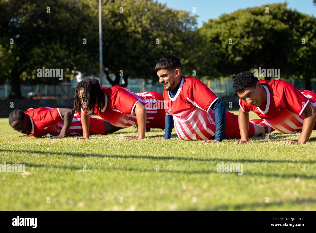 Des joueurs multiraciaux masculins portant des uniformes rouges font des push-up sur un terrain de jeu herbeux en été Banque D'Images