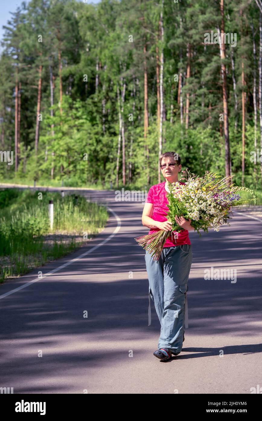 portrait complet d'une femme souriante heureuse avec un énorme bouquet de fleurs sauvages marchant le long d'une route forestière Banque D'Images