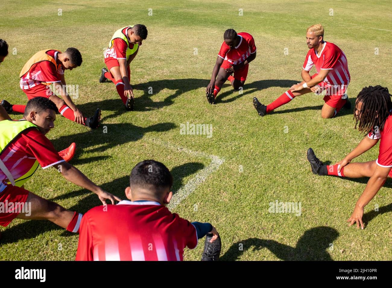Vue en grand angle des athlètes masculins multiraciaux qui s'étirent les jambes sur un terrain herbeux dans le terrain de jeu en été Banque D'Images