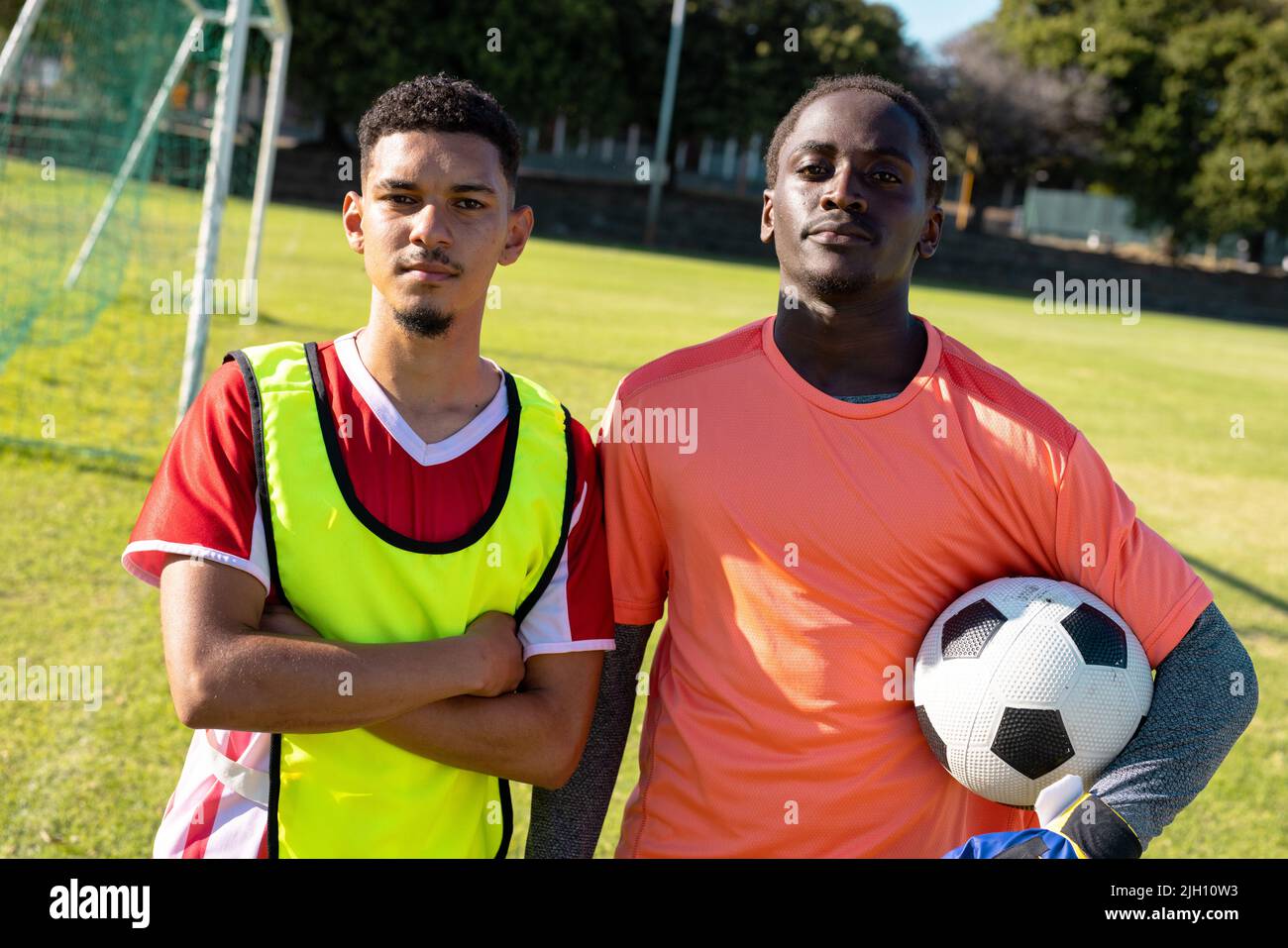 Portrait de joueurs multiraciaux sérieux avec les bras croisés et tenant le ballon debout dans le terrain de jeu Banque D'Images