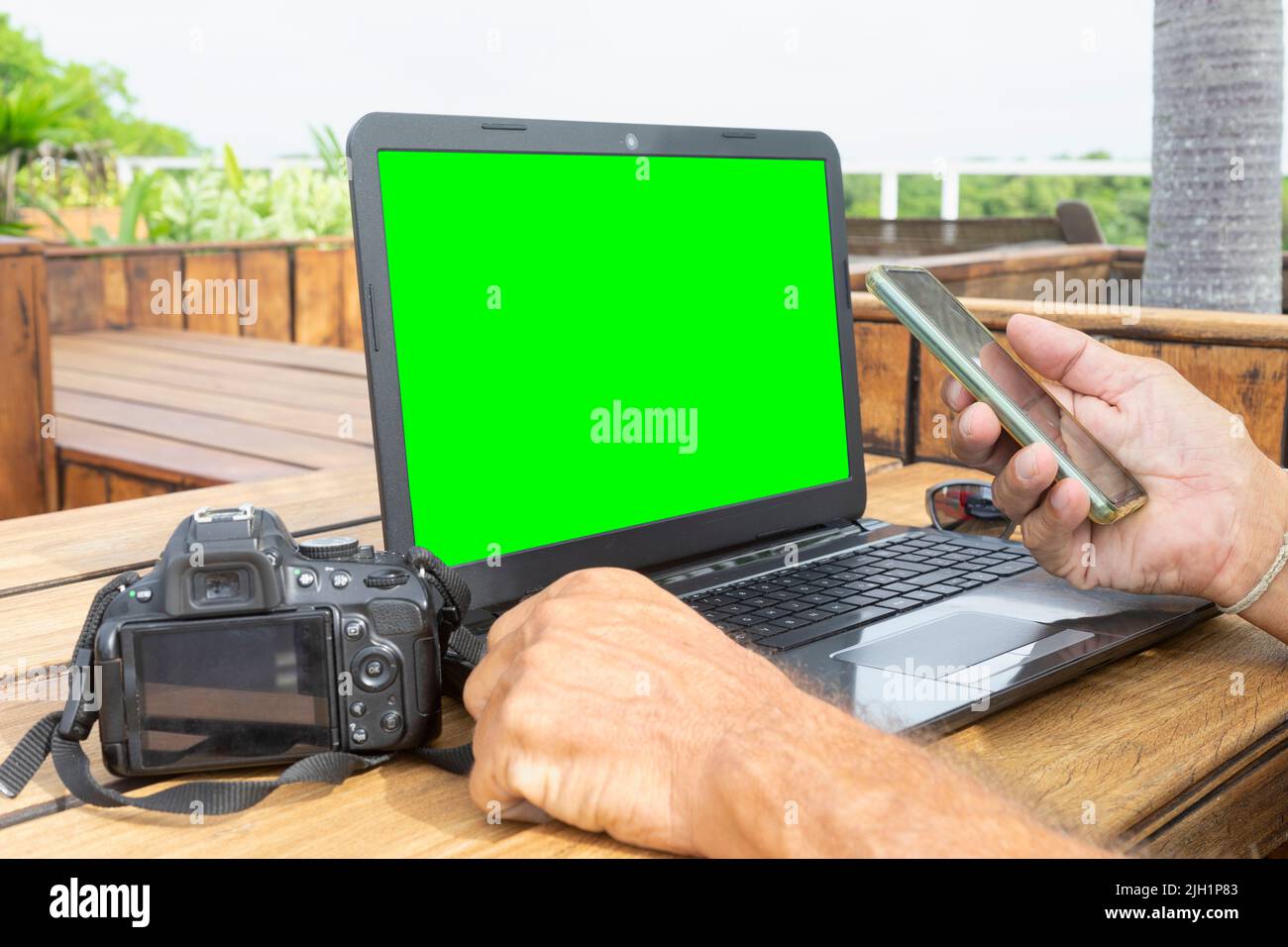 Gros plan d'un photographe freelance travaillant avec un smartphone, un ordinateur portable avec un écran vert appareil photo reflex vide sur une table en bois extérieure. Banque D'Images