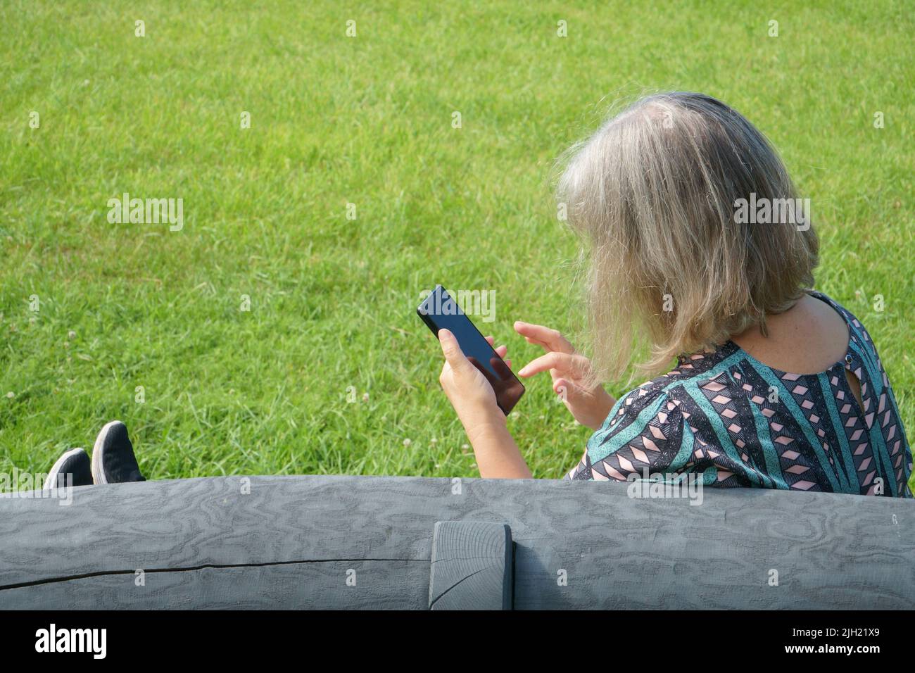 Une femme d'environ 60th ans en loisirs s'assoit sur le banc avec un smartphone dans sa main et cherche de nouveaux messages. Femme d'âge senior dans la nature pendant le Banque D'Images