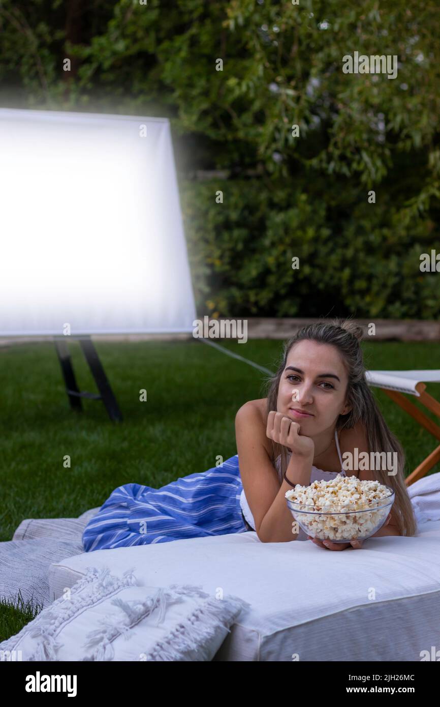 Vue verticale de la jeune belle femme regardant la caméra avec un bol complet de popcorns dans le cinéma en plein air dans le jardin la nuit Banque D'Images