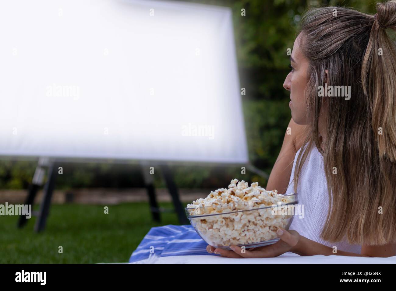 Vue latérale d'une femme blonde non reconnaissable regardant un film dans le jardin avec un projecteur et tenant des popcorns Banque D'Images