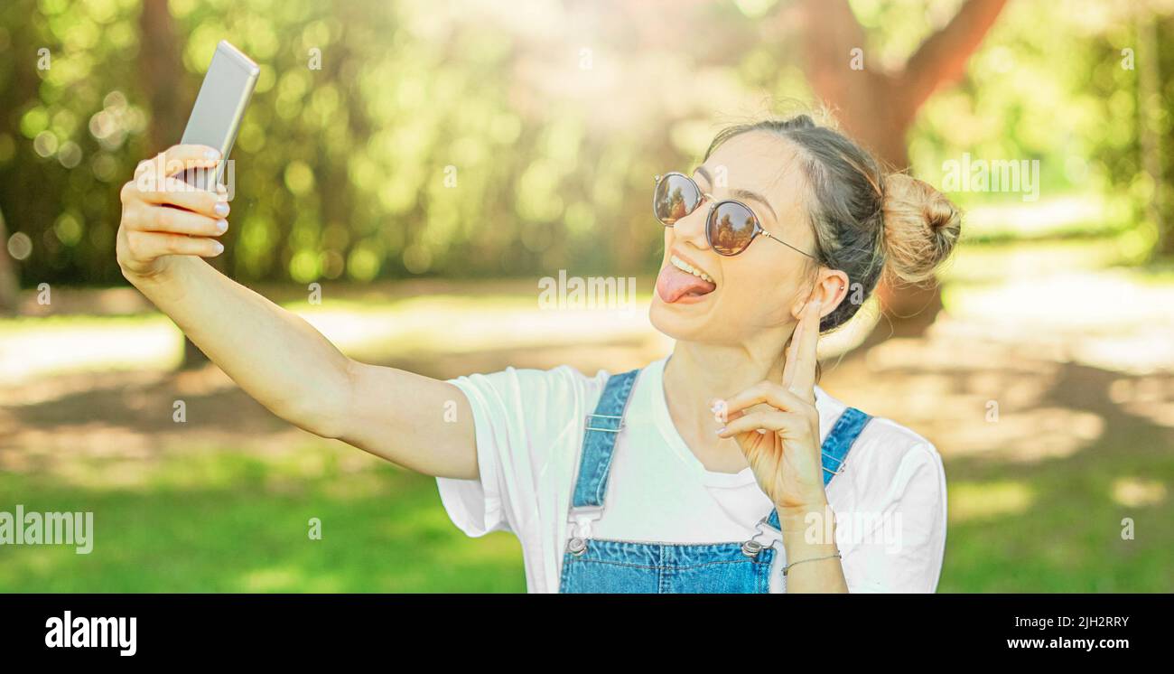 Bonne joyeuse jeune fille dans la nature prenant selfie avec espace coiffure de pain - Teeneger faisant le visage drôle et tenant son smartphone Banque D'Images