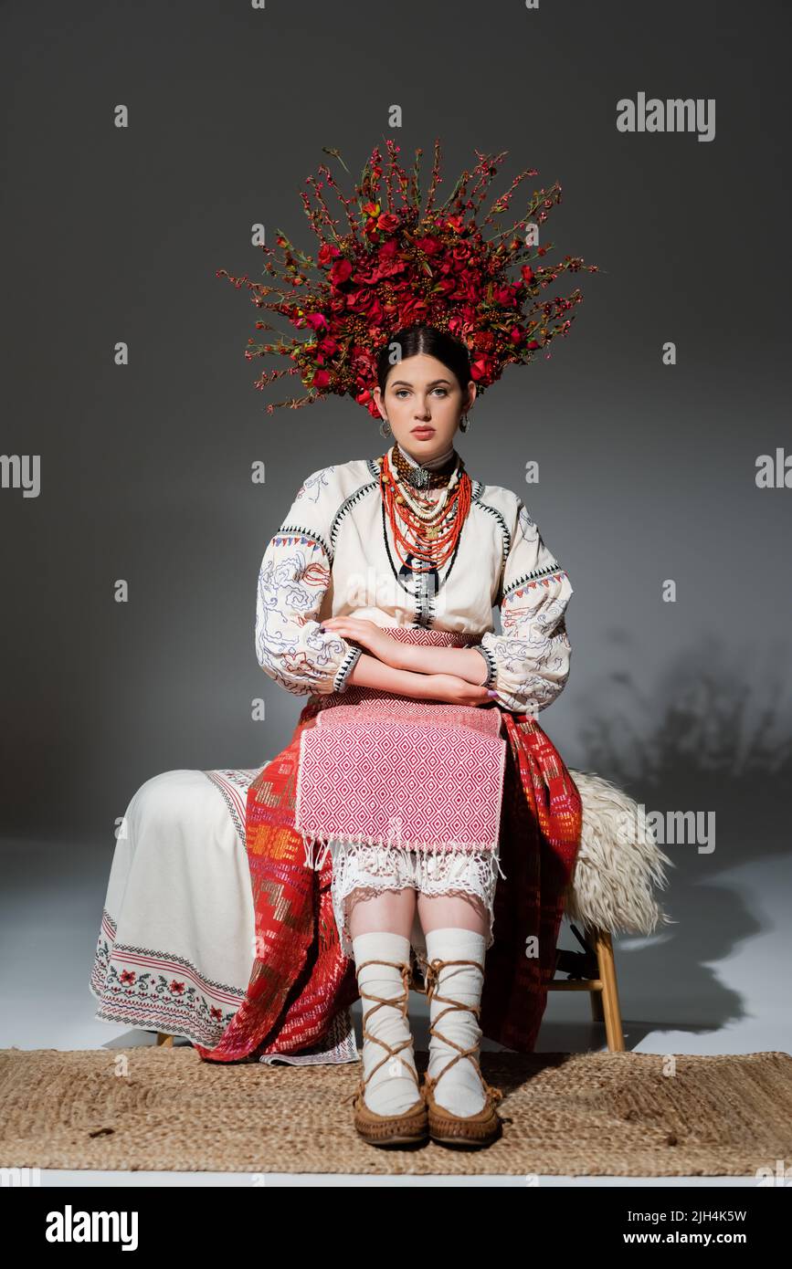 pleine longueur de jeune femme ukrainienne en vêtements traditionnels et couronne florale rouge assise sur un banc gris Banque D'Images