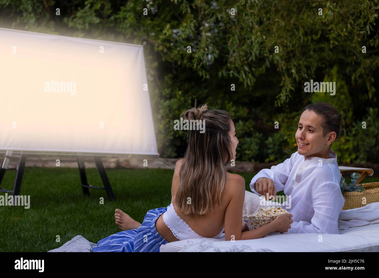 Un jeune couple heureux se regardant les uns les autres et regardant un film dans le cinéma d'été en plein air avec un bol de pop-corn dans le jardin Banque D'Images
